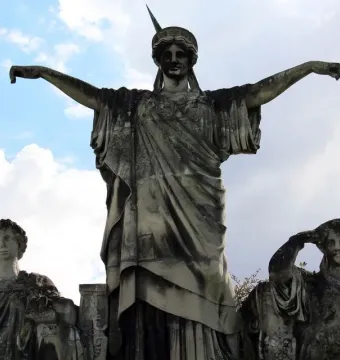 Weathered stone sculpture of France crowning Art and Industry, seen from below against a cloudy sky.