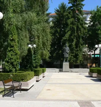Hristo Botev monument on a stone pedestal in a paved square, flanked by trees and hedges.