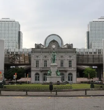 The John Cockerill Monument stands on a pedestal in a circular grassy area, with a historic building and modern glass towers behind it.