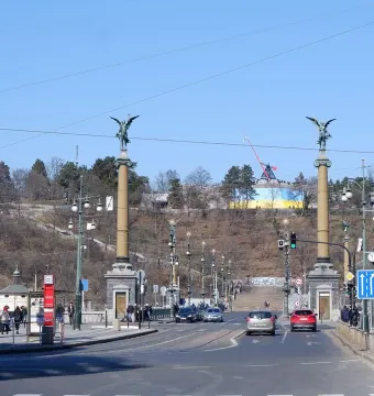Prague Metronome rising above a tree-lined hillside with a large yellow and blue banner below it.