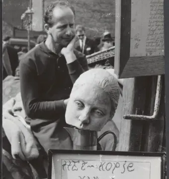 A sculpted female head resting on a metal pitcher, with a man in the background and a framed handwritten sign at right.