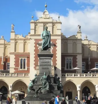 Adam Mickiewicz Monument in a paved square, with bronze figures on a tall pedestal and an ornate building behind it.