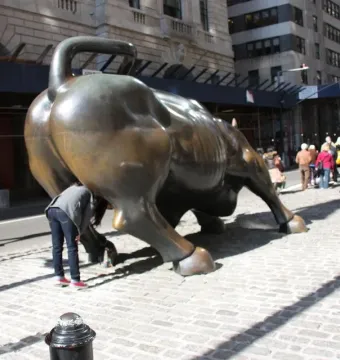 Charging Bull bronze sculpture seen from the rear on a cobblestone street with pedestrians nearby.