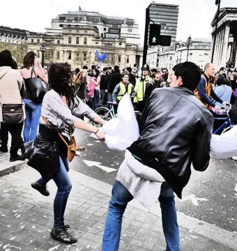 A man and woman in a lively pillow fight in a city plaza, with a crowd and buildings behind them.