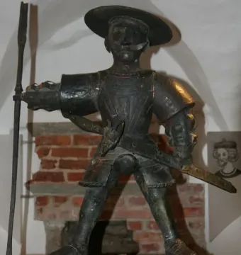 Old Thomas statue in armor with a wide-brimmed hat, mustache, sword, and long pole-like weapon.