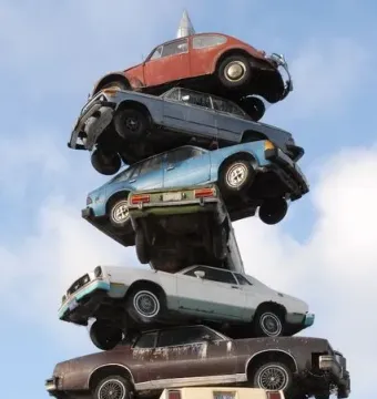 Spindle sculpture with old cars stacked vertically on a central pole, topped by a red Volkswagen Beetle against a blue sky.