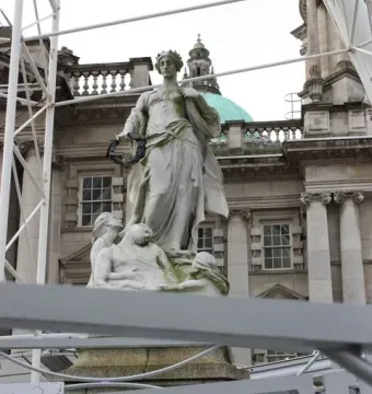 Titanic Memorial statue in Belfast, with a crowned woman holding a wreath above smaller figures and a building with a green domed roof behind it.
