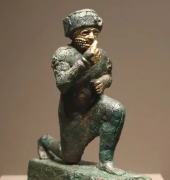 Worshipper of Larsa, a kneeling ancient metal figure with a raised hand, gold-plated face and hands, and a small bowl on its base.