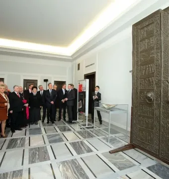 Replica of the Gniezno Doors displayed upright on wooden supports in a bright room with visitors nearby.