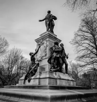 The Marquis de Lafayette statue rises on a multi-tiered pedestal with smaller figures at the base, photographed in black and white against bare trees and an overcast sky.