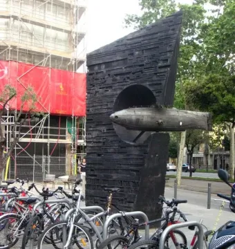 Monument to Narcís Monturiol with a scale submarine model emerging from a dark concrete base, with bicycles in front and a scaffolding-covered building behind.