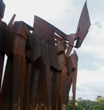 The Monument to the Azoreans, a tall rusted metal sculpture with white graffiti near the base, stands on a grassy area beneath a cloudy sky.
