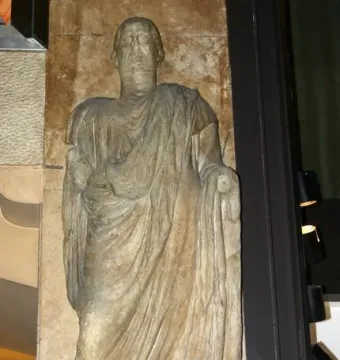 Weathered stone statue of Scior Carera standing on an inscribed base against a stone wall.