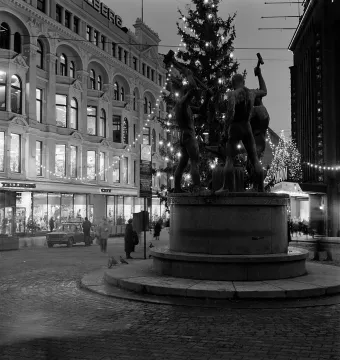 The Three Smiths statue with three muscular figures holding hammers around an anvil in a city square, with a lit Christmas tree behind it.