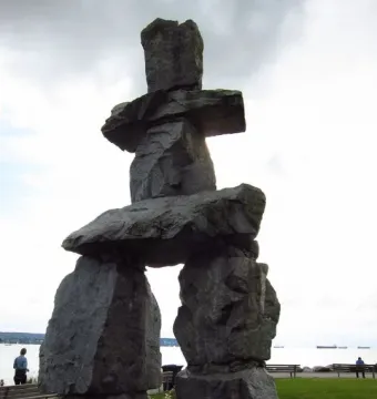 An Inukshuk stone sculpture stands on a grassy waterfront lawn under an overcast sky.