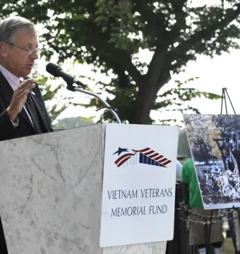 A man speaks at a podium beside a Vietnam Veterans Memorial Fund sign, with a framed display of The Three Soldiers statue on an easel decorated with flowers and flags.