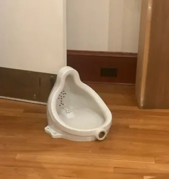 A white glazed ceramic urinal sits on a polished wooden floor, with dark dots visible inside and a circular drain opening near the base.