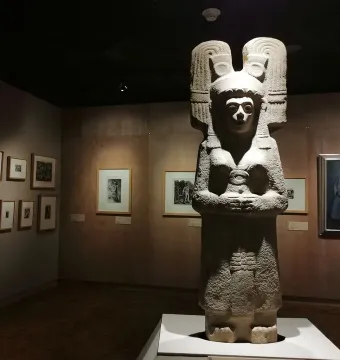 The Young Woman of Amajac stone statue standing in a museum gallery with an ornate headdress and clasped hands.