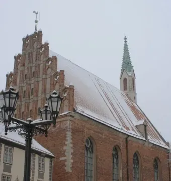 A winter scene with a red-brick building, a green spire, a streetlamp, and people walking near a snowy wall.