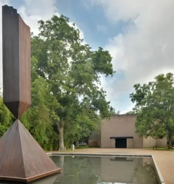 Barnett Newman’s Broken Obelisk sculpture rising from a reflecting pool with trees and a light-toned building behind it.