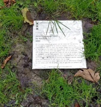 A rectangular plaque for De Schreeuw lies in damp ground among grass and scattered leaves.