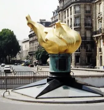 The Flame of Liberty sculpture with a golden flame on a dark stepped base in an urban street setting.