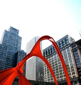 Flamingo, a large red abstract steel sculpture, stands in a city plaza with tall buildings in the background and a person near its base.