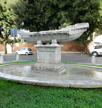 Fontana della Navicella, a weathered stone boat-shaped fountain in an oval basin surrounded by grass and trees.