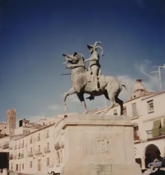 Francisco Pizarro equestrian statue on a stone pedestal in a town square, with historic buildings and a tower behind it.