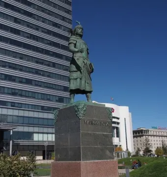 Marco Polo statue in historical attire holding a book on a stone pedestal.