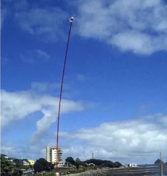 The Wind Wand sculpture rises from a coastal promenade beside the shoreline and city buildings under a blue sky.