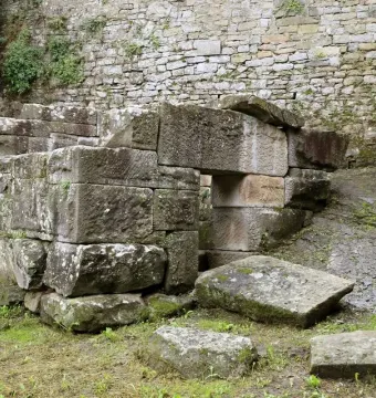 Etruscan tomb built of weathered stone blocks with a small square entrance, set beside a stone wall.