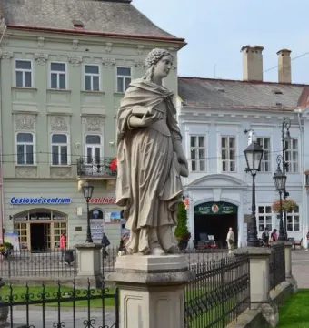A stone Marian column figure stands behind a wrought-iron fence on a city street.