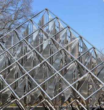 Delta Solar pyramid sculpture made of a metallic grid with reflective triangular panels, set against bare trees and a light-colored building.