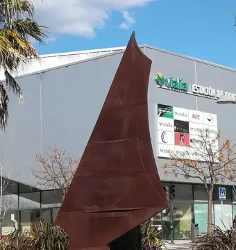 Dorna, a rust-colored abstract metal sculpture with a pointed top and splayed base, set on a stone wall among shrubs.