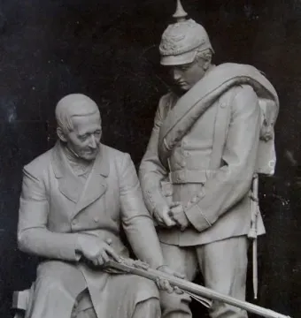 Dreyse Monument sculpture showing a seated man examining a rifle beside a soldier in a spiked helmet.