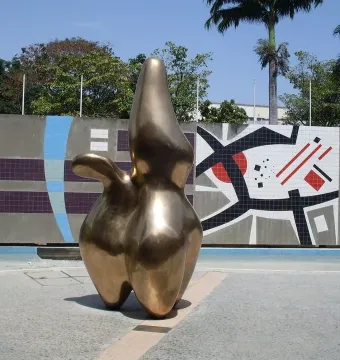 Cloud Shepherd, a smooth golden sculpture, stands on a paved plaza beside a geometric tiled wall.