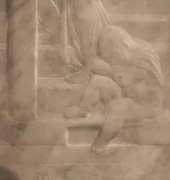 Close-up of a small sculpted figure from Donatello\'s Feast of Herod, seated on steps beside draped fabric.
