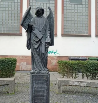 Frankfurter Engel, a dark winged angel statue holding a scroll and a book on a pedestal in a cobblestone plaza.