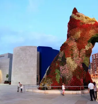 The Puppy flower sculpture stands in an outdoor plaza, covered in red, pink, white, and green flowers, with people gathered nearby.