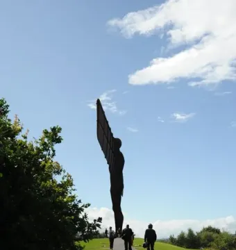 The Angel of the North sculpture stands on a grassy hill beside a paved path with people walking toward it under a blue sky.