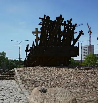 Monument to the Fallen and Murdered in the East, a dark sculpture of crosses and wooden beams on a raised cobblestone platform, with an inscribed stone in front.