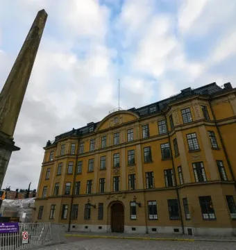 The Obelisk at Slottsbacken stands beside a large yellow building and metal fence under a cloudy sky.