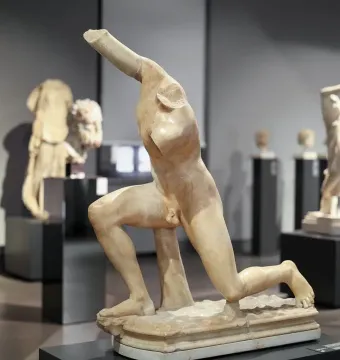 Subiaco Ephebe, a pale stone kneeling male sculpture displayed on a dark pedestal in a museum gallery.
