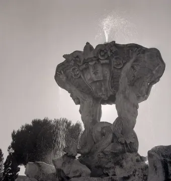 The Fountain of the Tritons with two muscular figures supporting a shell-shaped basin as water sprays upward and cascades down.
