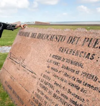 A woman stands beside the Holocaust Memorial plaque, a large angled pink granite monument with engraved text, near a grassy waterfront.