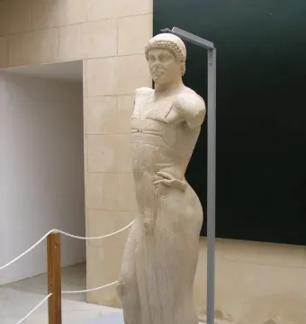 The Motya Charioteer statue displayed on a gray platform in a museum hall.