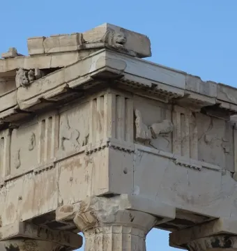 Weathered marble corner of the Parthenon pediments with fluted columns, triglyphs, worn relief metopes, and a lion\'s head against a blue sky.