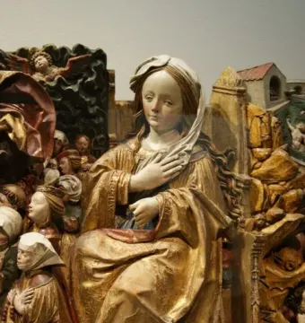 A carved wooden seated woman in a gold robe and white headdress, surrounded by smaller figures and rocky architectural details.