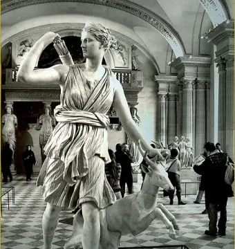 The Diana of Versailles marble statue stands in a museum hall with a small deer beside it and visitors in the background.